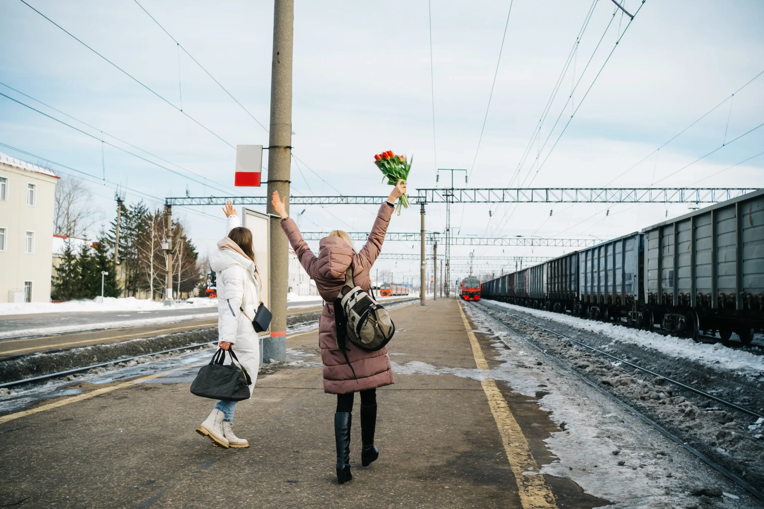 two girls at the station happily with flowers meet a train that's coming towards the station